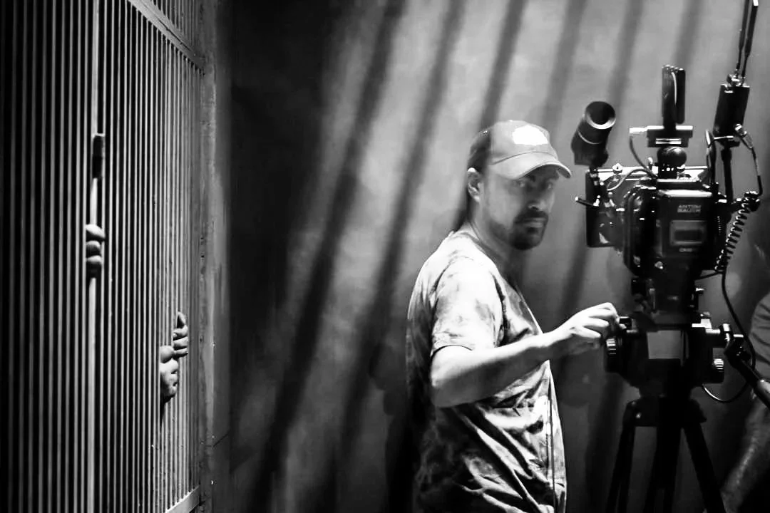 A man operating a film camera in a dimly lit studio with curtains and a person peeking through a vent on the left side.