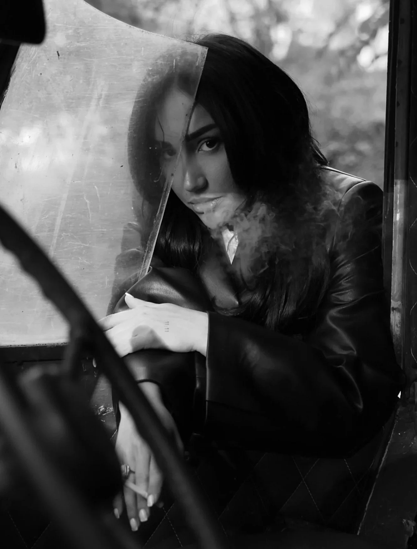 A woman with long dark hair and dark eye makeup looking through a small glass partition, with visible smoke and indoor background.