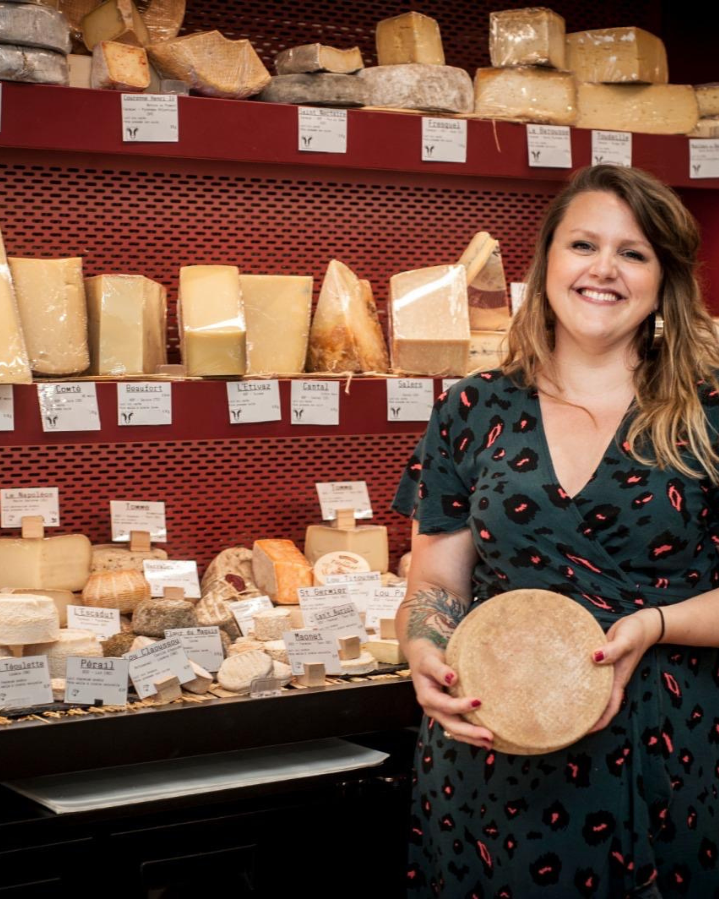 Femme souriante tenant un fromage rond dans une fromagerie avec plusieurs fromages sur les étagères derrière elle.