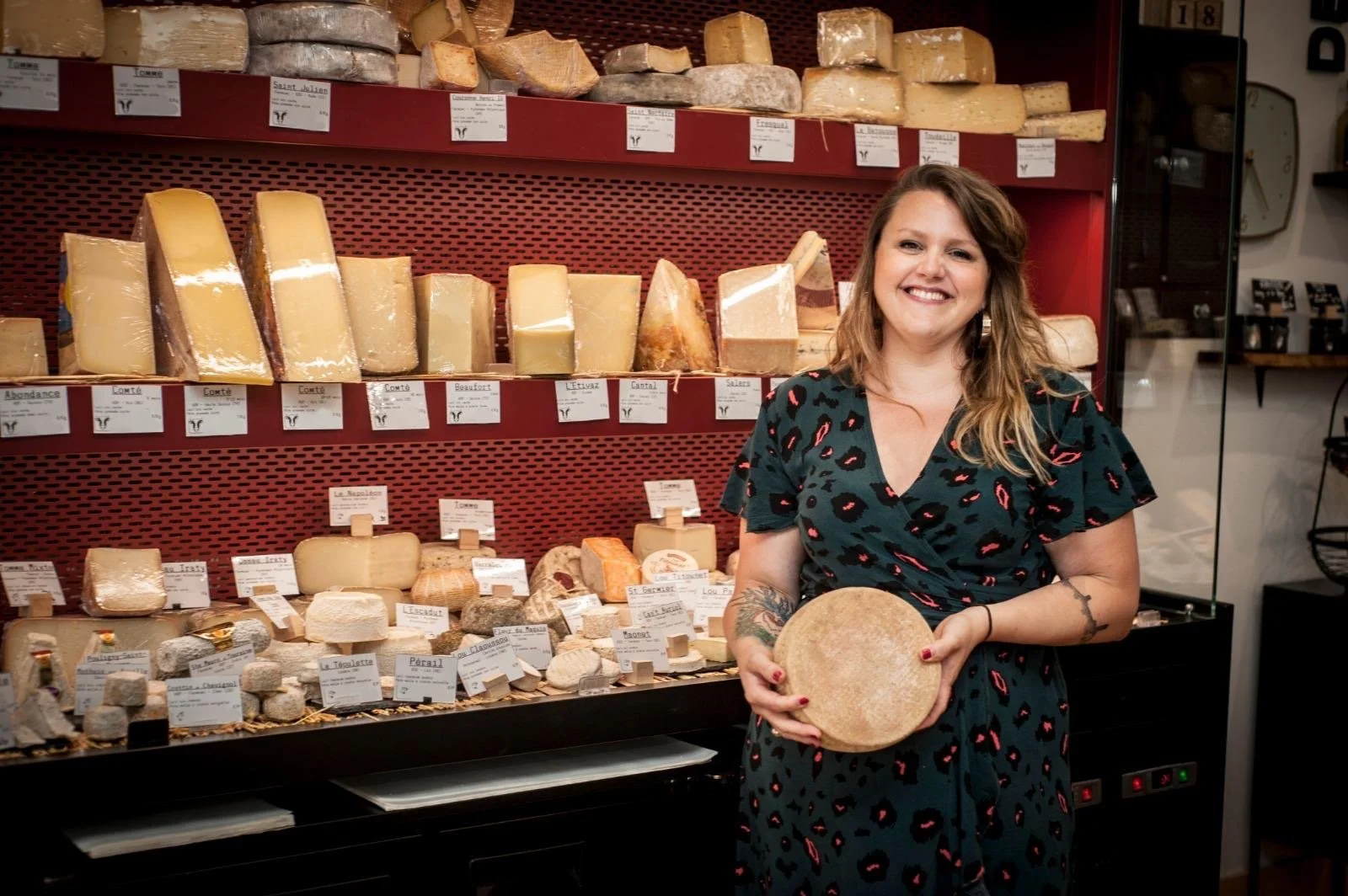 Femme souriante tenant un fromage rond dans une fromagerie, avec un étalage varié de fromages en arrière-plan.