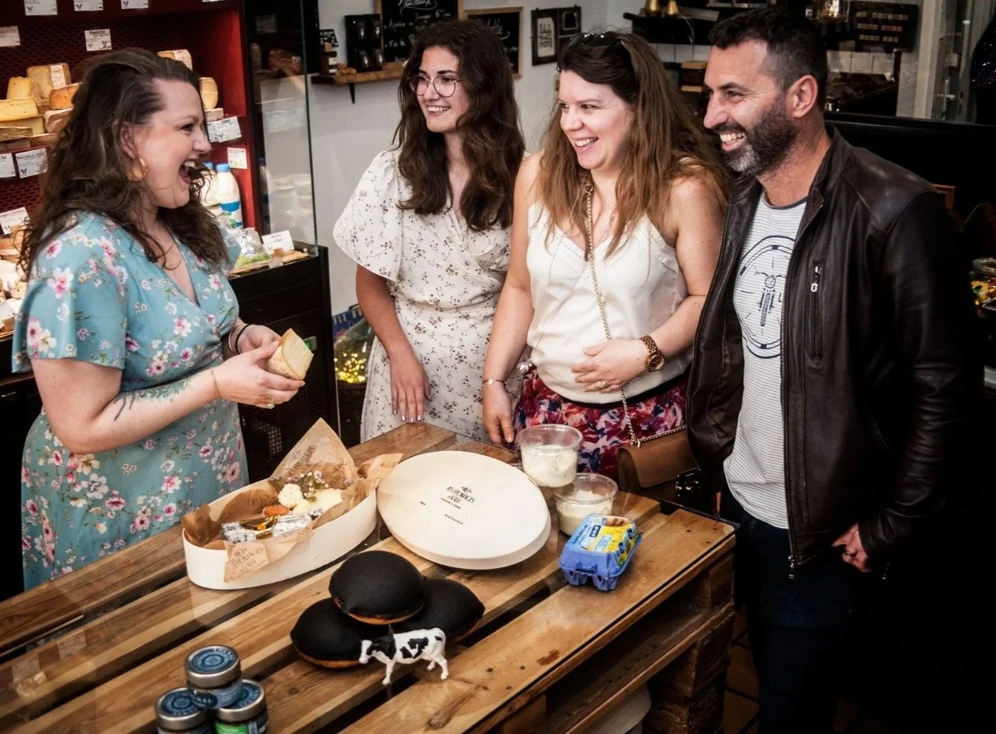 Quatre personnes souriantes autour d'une table à l'intérieur d'une épicerie, où une femme en robe bleue tient un morceau de pain, avec un gâteau au chocolat, des œufs, et d'autres aliments sur la table.