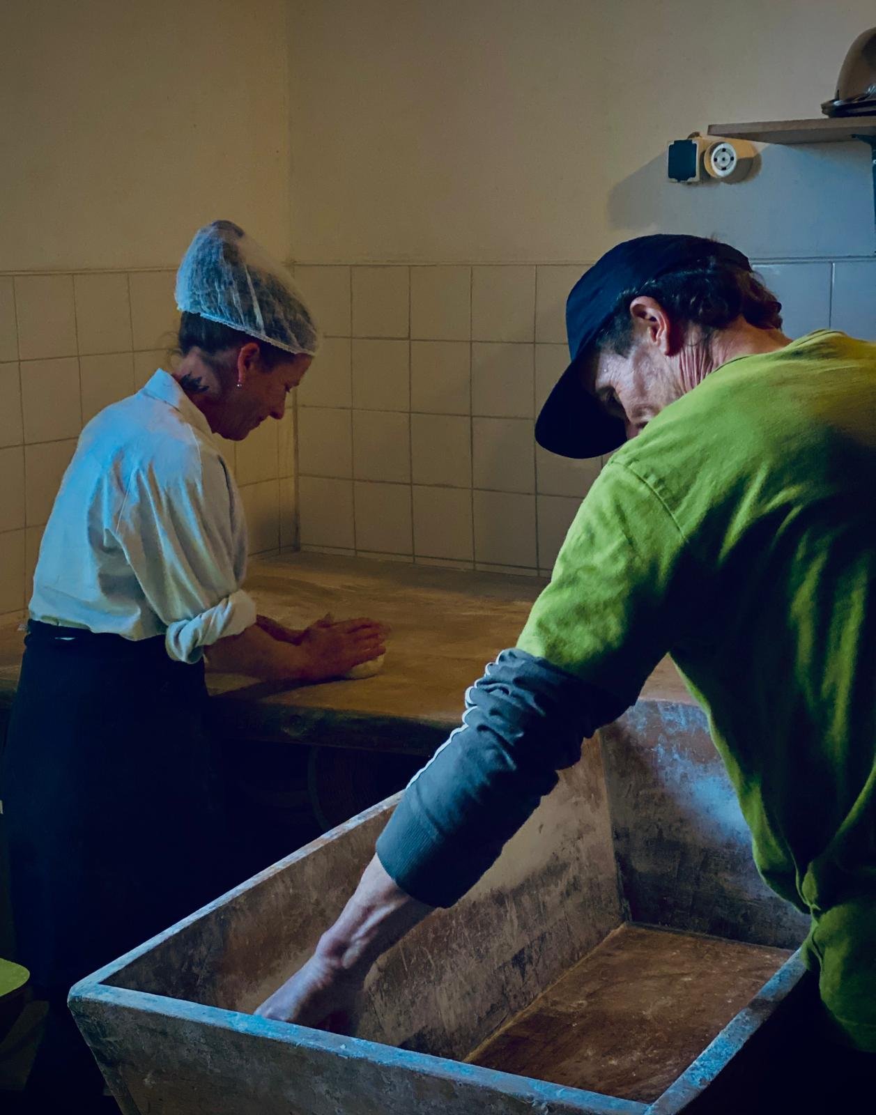 Deux personnes travaillent dans une cuisine, l'une mélange une pâte sur un plan de travail en bois, l'autre plonge sa main dans un grand récipient en métal.