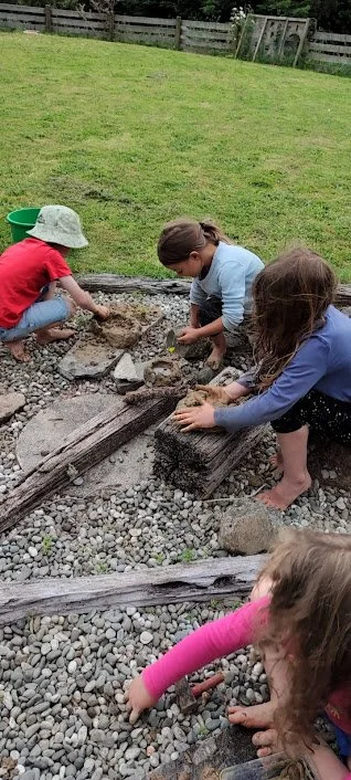 Three children working together on a gravel and wooden railway track, with grass and a wooden fence in the background.