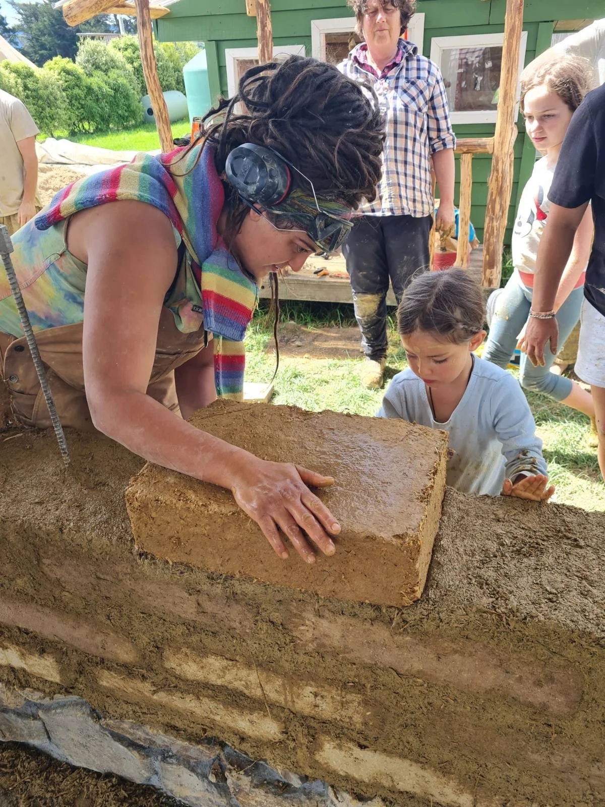 A woman with glasses and a colorful scarf working on an adobe brick in a construction site with a small group of people observing, including a young girl in a white shirt and others standing outdoors with trees and a green building in the background.