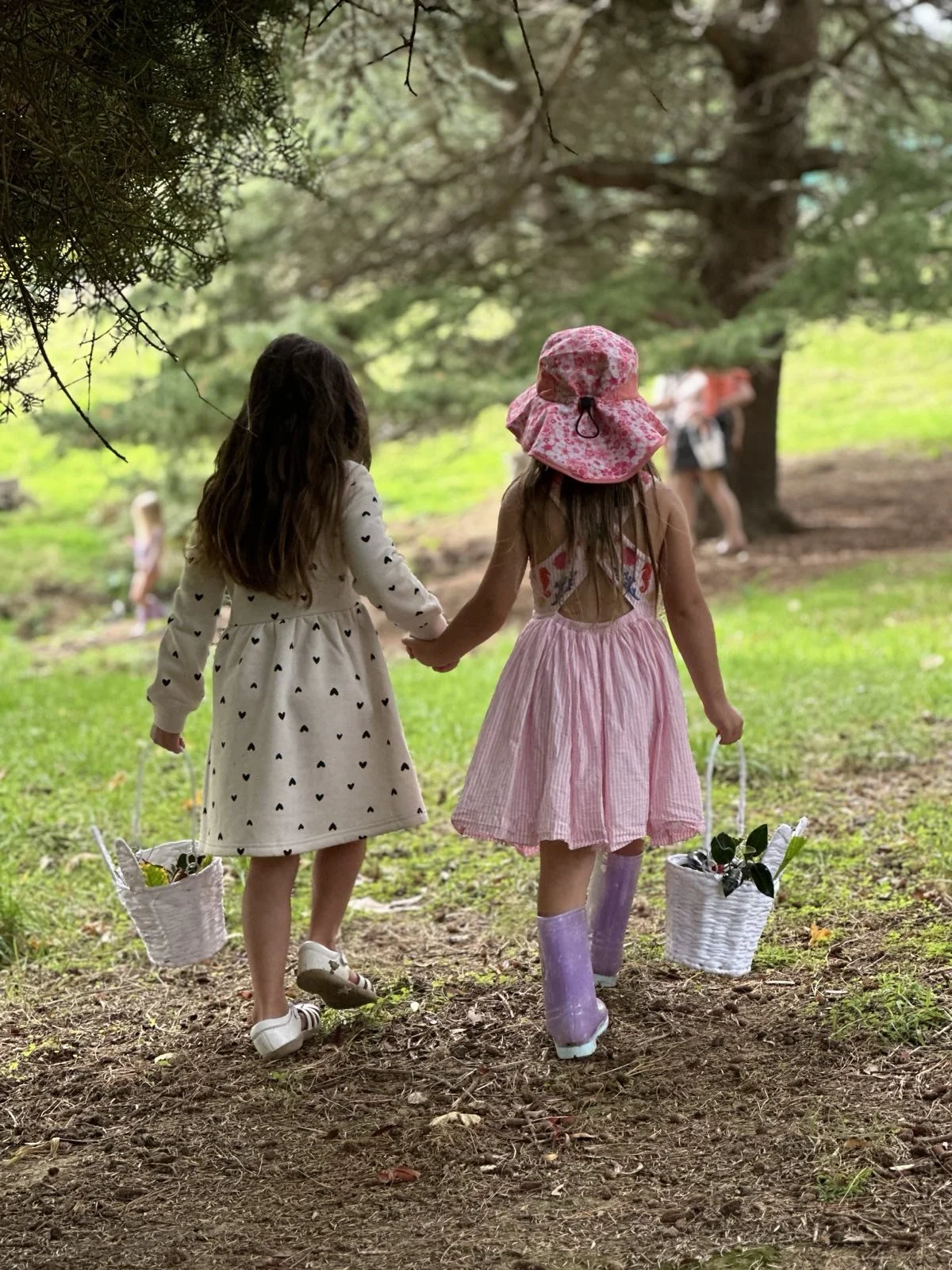 Two young girls walking hand-in-hand in a wooded park, carrying baskets with flowers. One girl wears a white dress with black hearts, the other a pink dress and a pink sun hat.