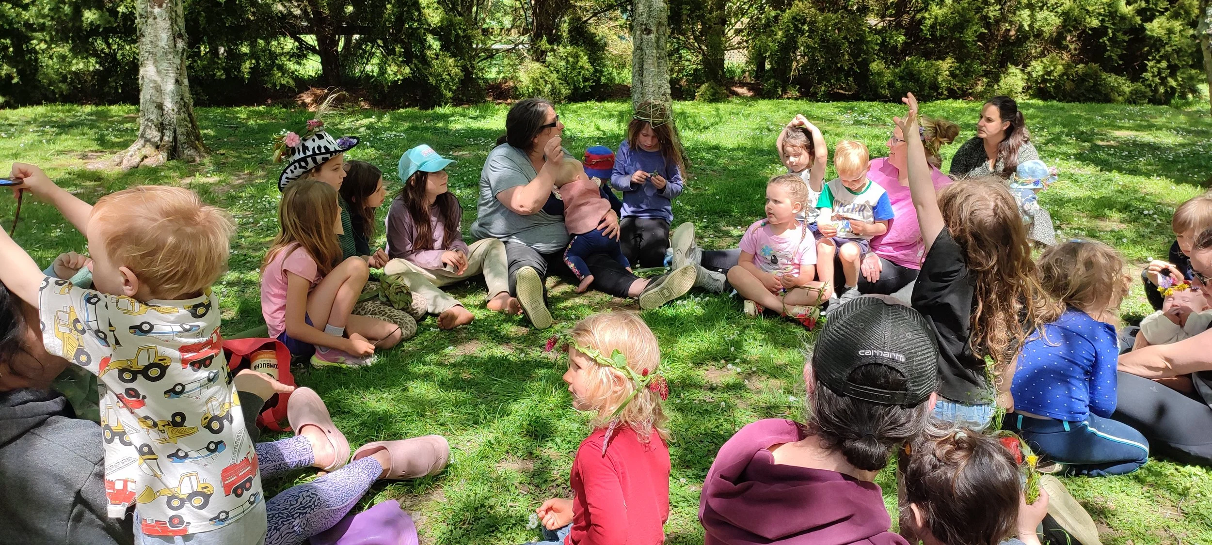 Children and adults sitting on grass in a park, engaging in an outdoor activity or storytelling session; some children wearing hats and raising hands, surrounded by trees and greenery.