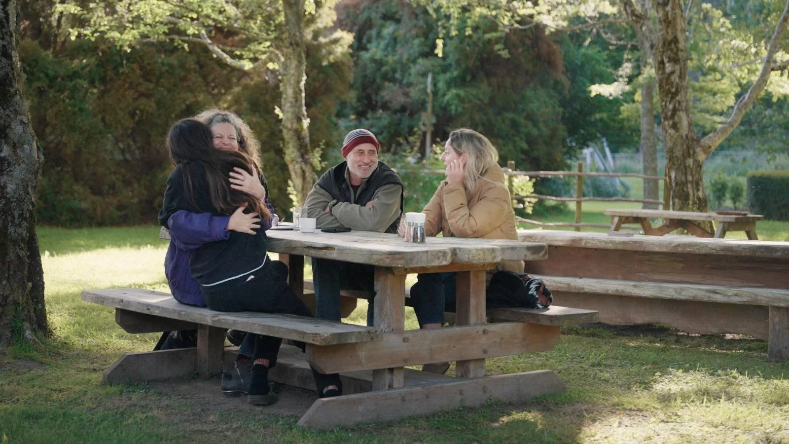 Four people sit at a wooden picnic table outdoors, with a woman hugging a child, while two others smile and watch. There are trees and grass in the background.