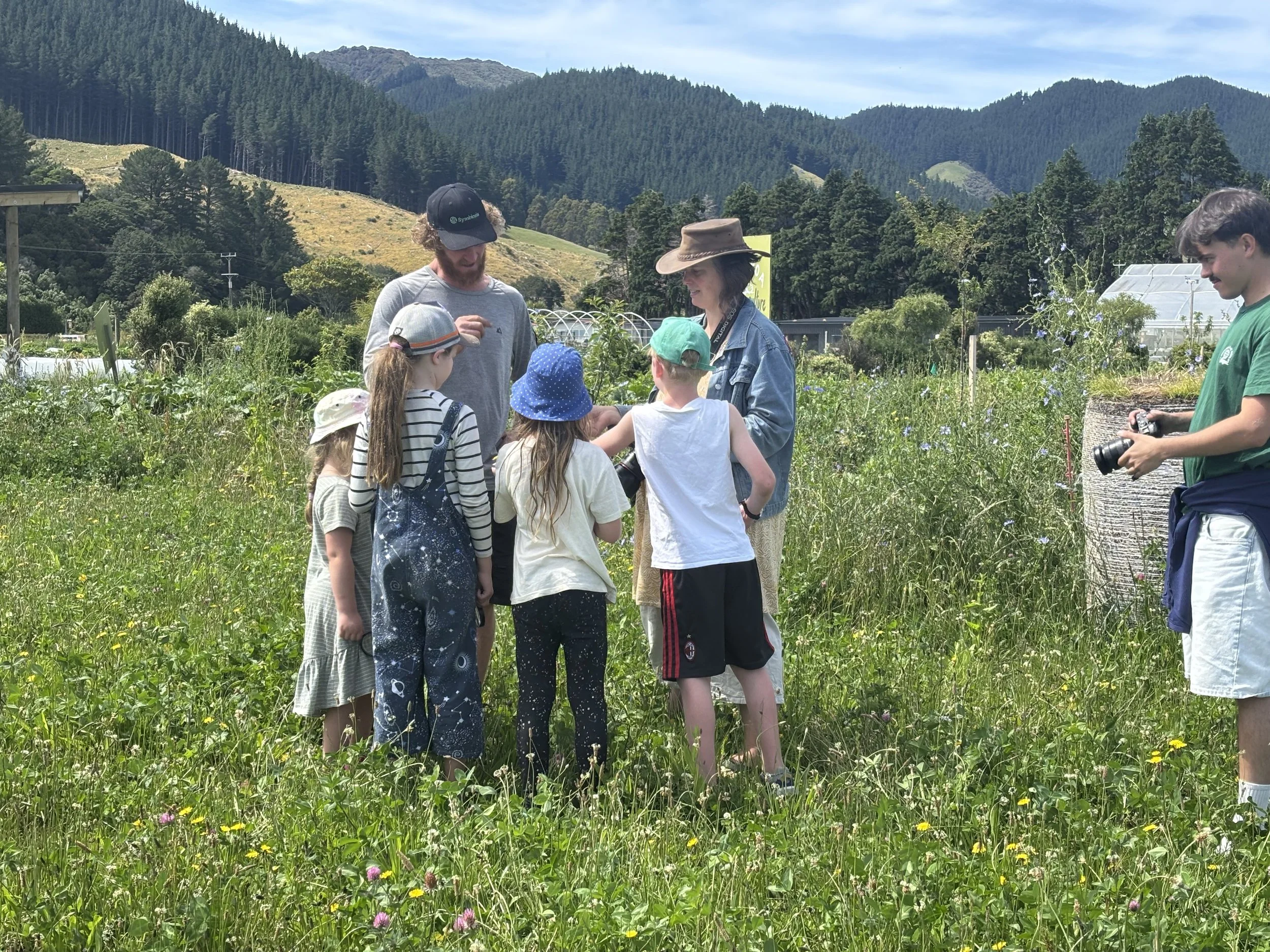 Children and adults in a lush green polyculture field with mountains in the background, engaging in an outdoor learning activity at Mangaroa Farms.