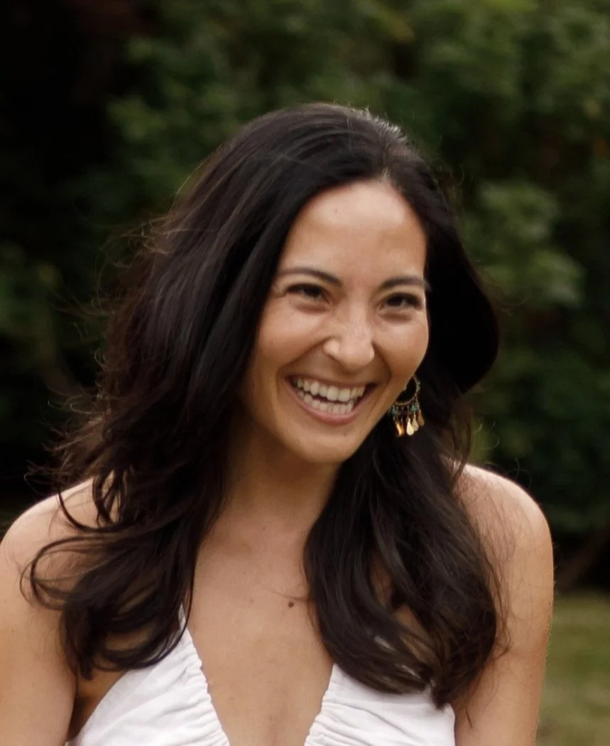 A woman with long dark hair smiling outdoors with a blurry green background.