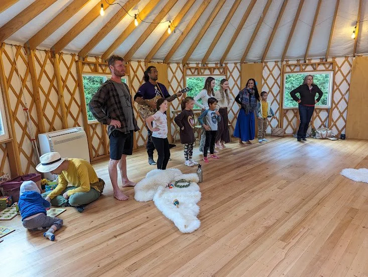 Group of children and adults in a yurt, with people standing and sitting, one person playing guitar, children singing or performing as they practice te reo Māori and waiata. cozy lighting.