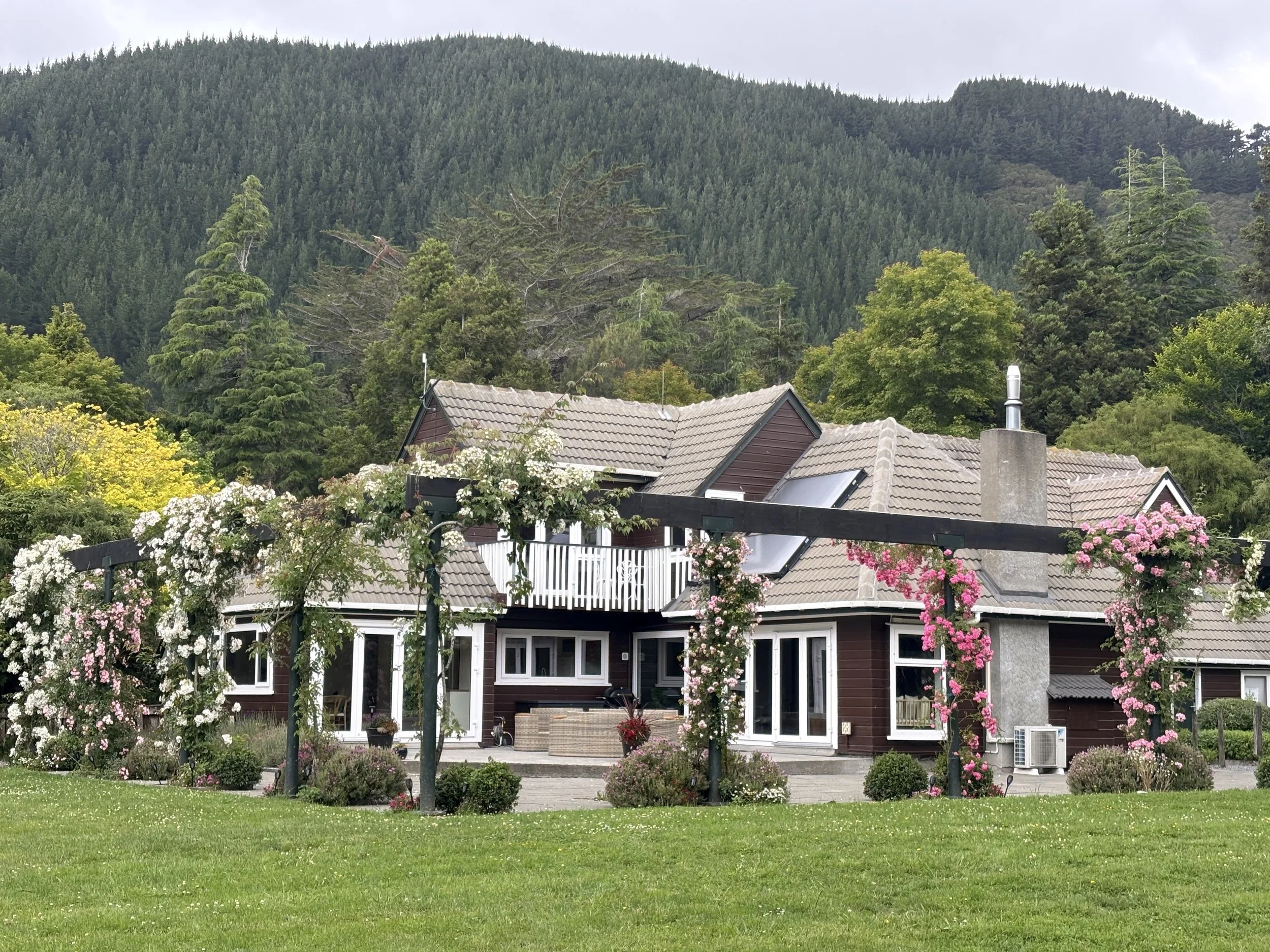 Woodcote Lodge at Mangaroa Farms. Worldschooling acommodation. A house with a porch and large windows, surrounded by flowering bushes with pink and white flowers, set against a backdrop of green hills with trees.
