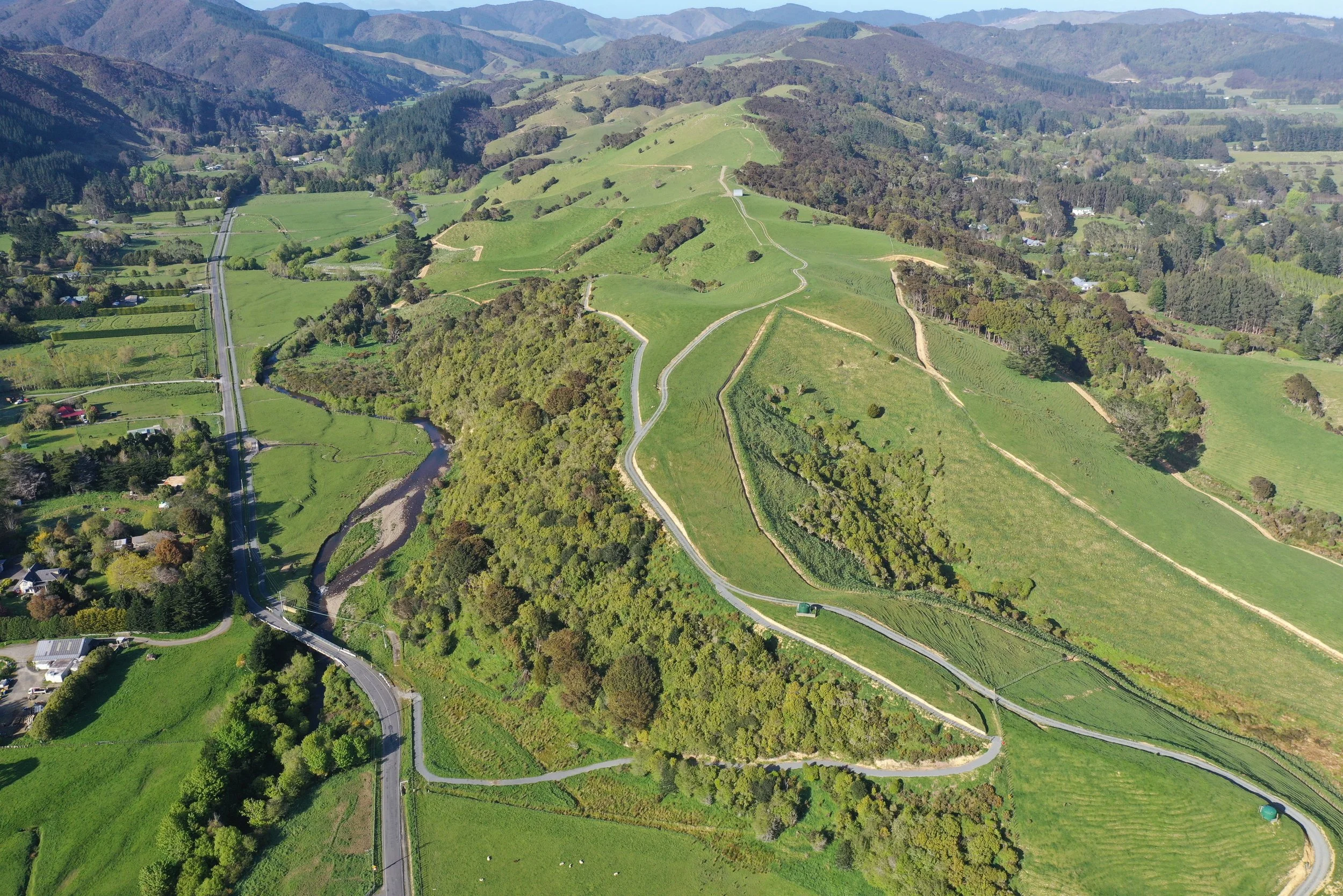 Aerial view of Mangaroa Farms' green rolling hills, winding dirt roads, a small stream, and scattered trees in a rural landscape.