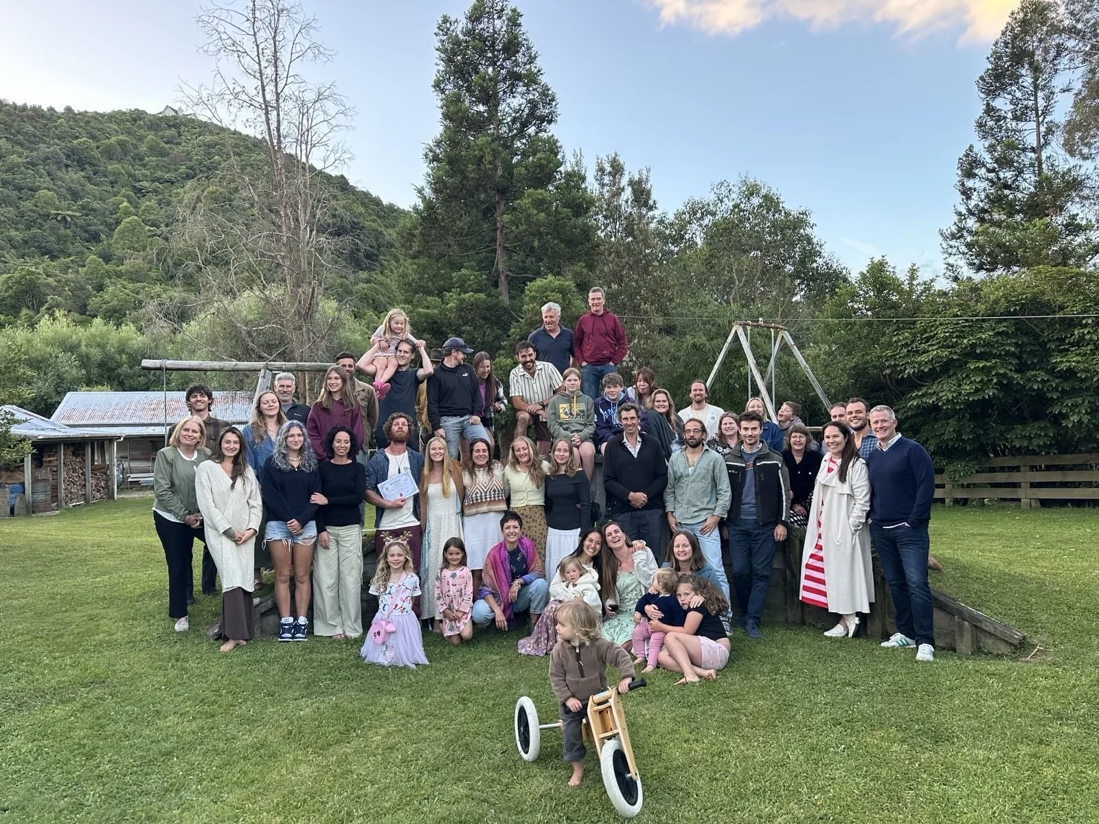A large group of people, including children and adults, gathered outdoors on a grassy area with trees and hills in the background. Some are smiling and posing, and a child is riding a small bicycle in the foreground. Mangaroa Farms team and community