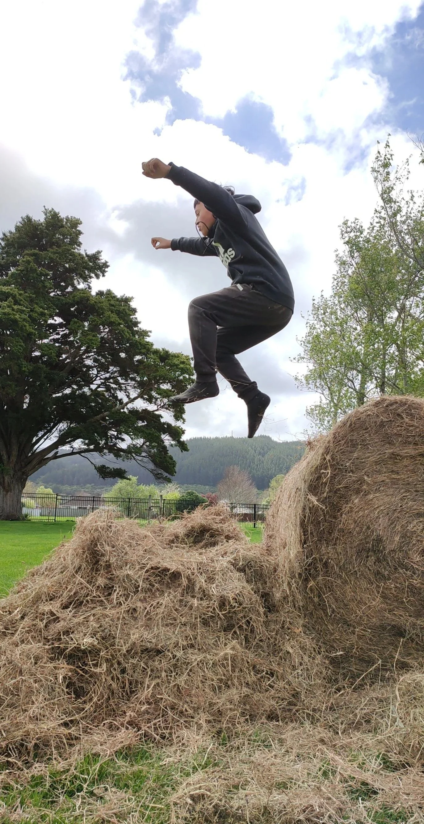 Child jumping off a pile of hay bales outdoors during daytime, with trees and cloudy sky in the background.