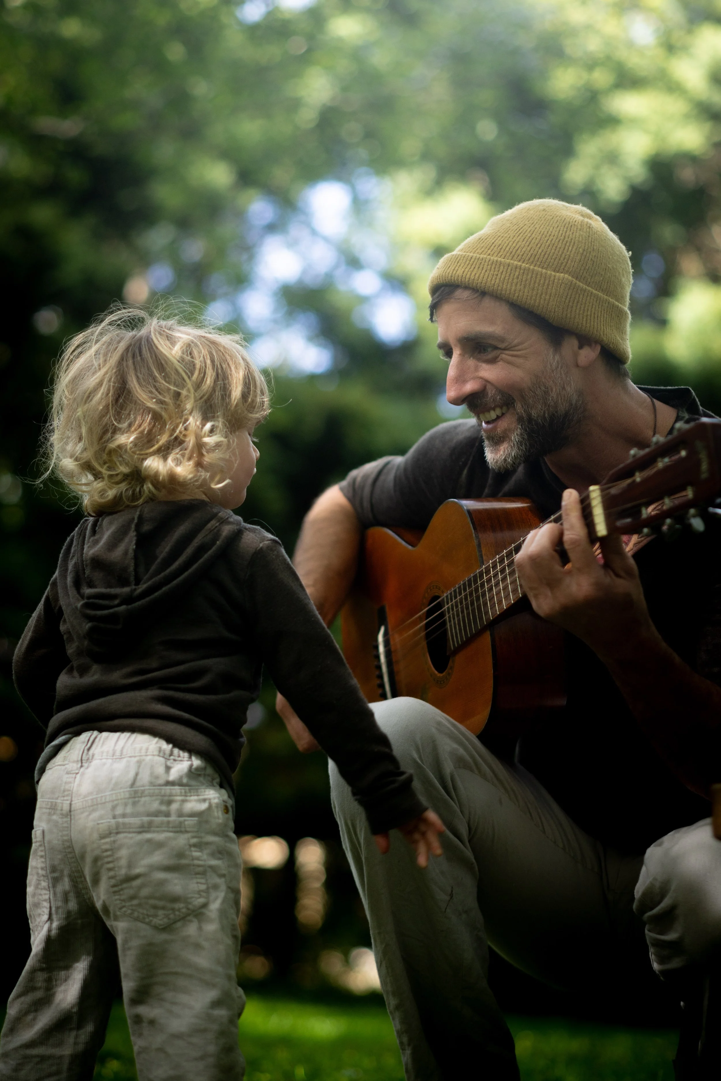A man with a beard wearing a yellow beanie playing guitar and smiling at a young child with curly blonde hair in an outdoor park setting.