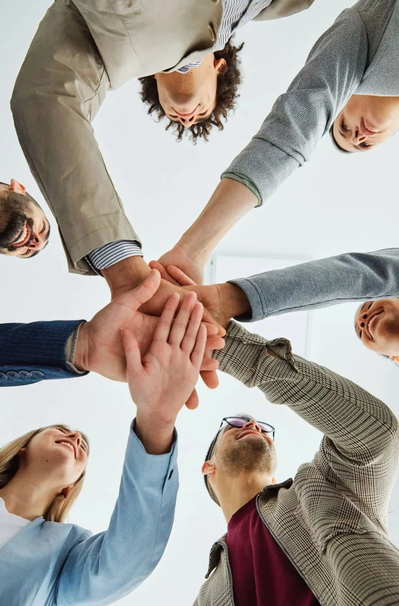 Group of diverse people in business attire putting their hands together in the center as a team, smiling and looking up.