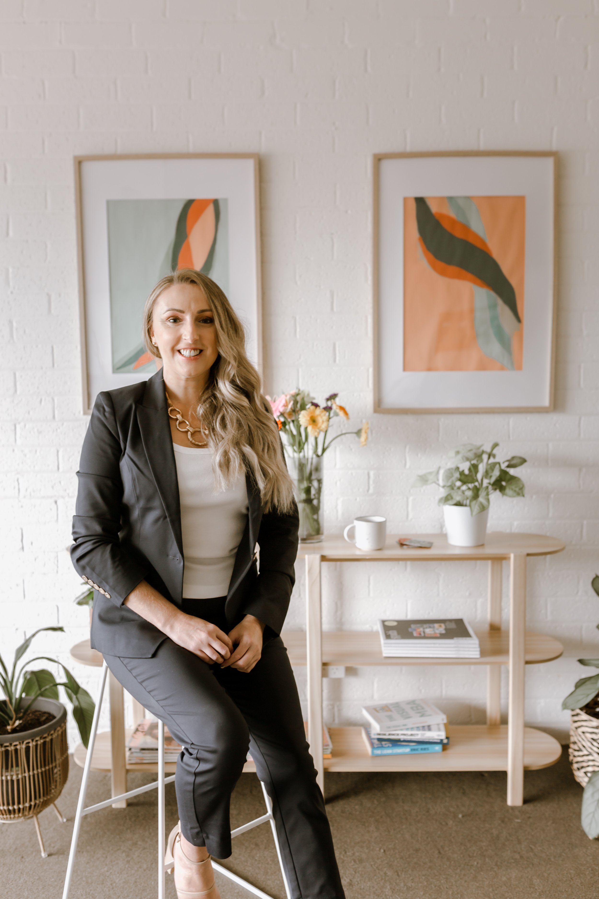 A woman with long blonde hair wearing a blue blazer and blue pants, sitting on a tall stool in a modern, decorated room with abstract art, a wooden bookshelf, and plants.