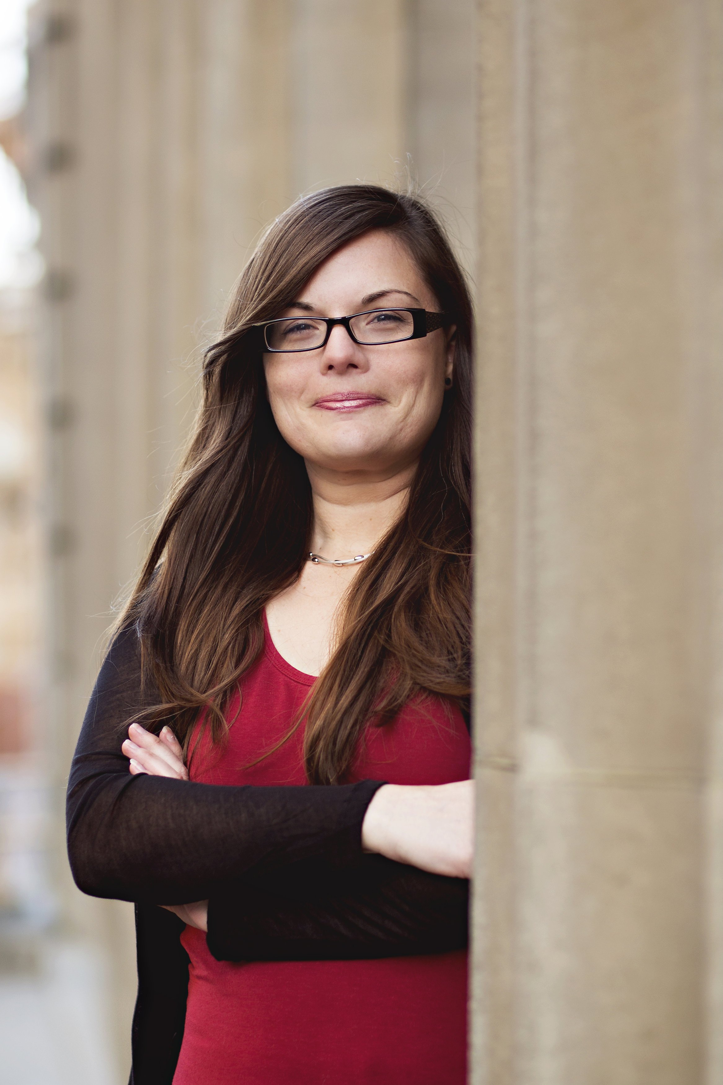 Young woman with dark brown hair and glasses wearing a red shirt and black overshirt standing behind a column.