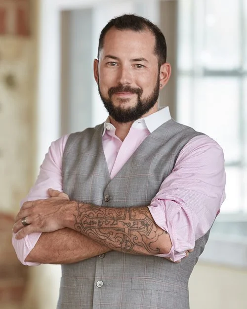 A confident man with a beard, tattooed arm, wearing a gray vest over a pink shirt, standing with crossed arms in a bright room.
