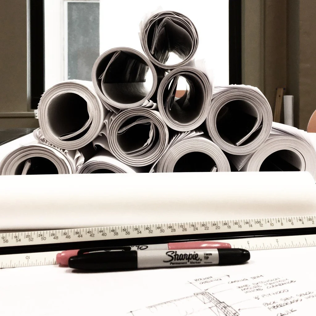 Several rolled-up architectural plans or blueprints stacked on a table, with a long ruler, a black and pink Sharpie marker, and handwritten notes and sketches in the foreground.