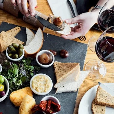 Person spreading pâté on bread at a charcuterie board with olives, salad, and glasses of red wine.
