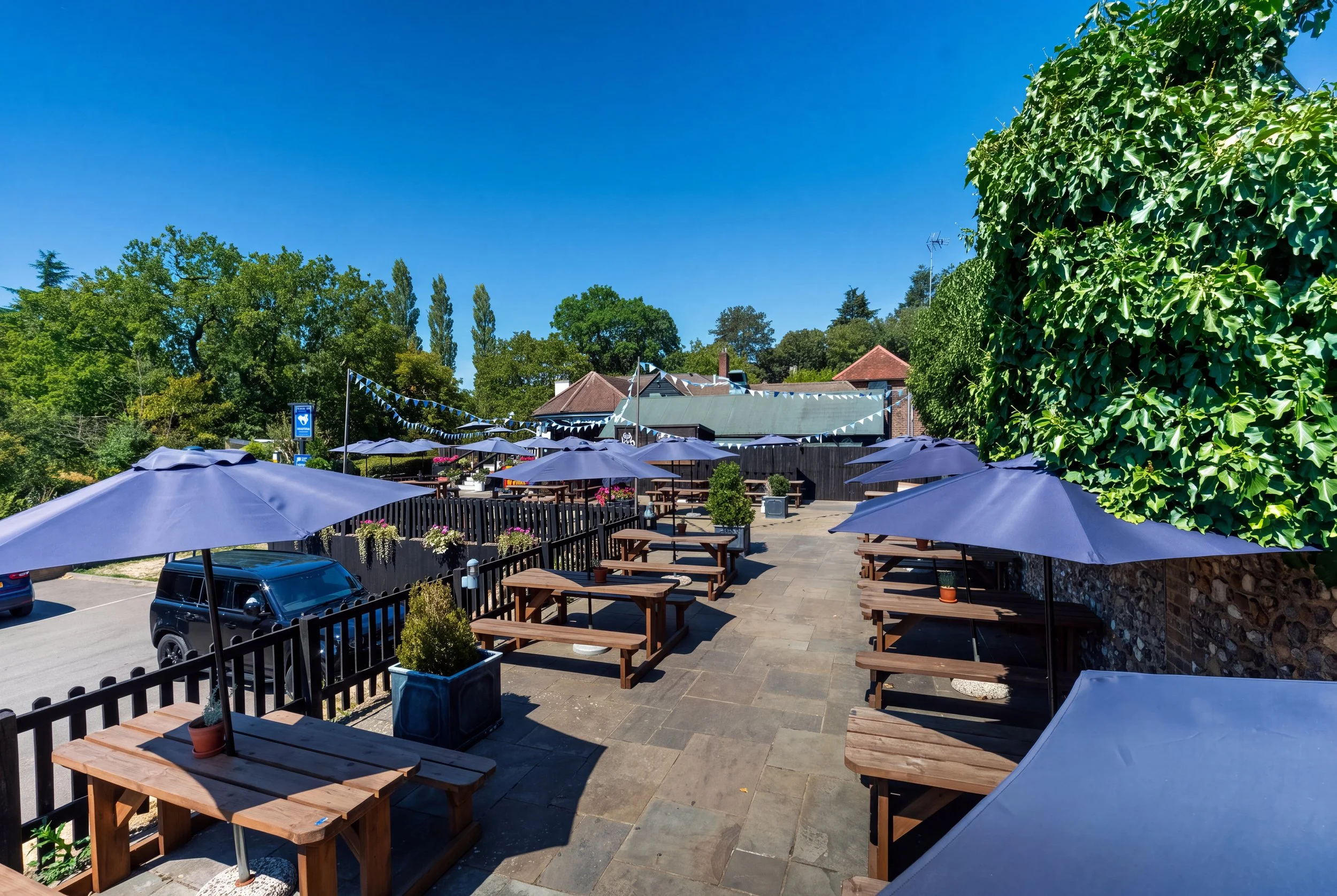 Outdoor patio area with wooden tables, purple umbrellas, and potted plants, surrounded by trees and a building with pennant banners under a clear blue sky.