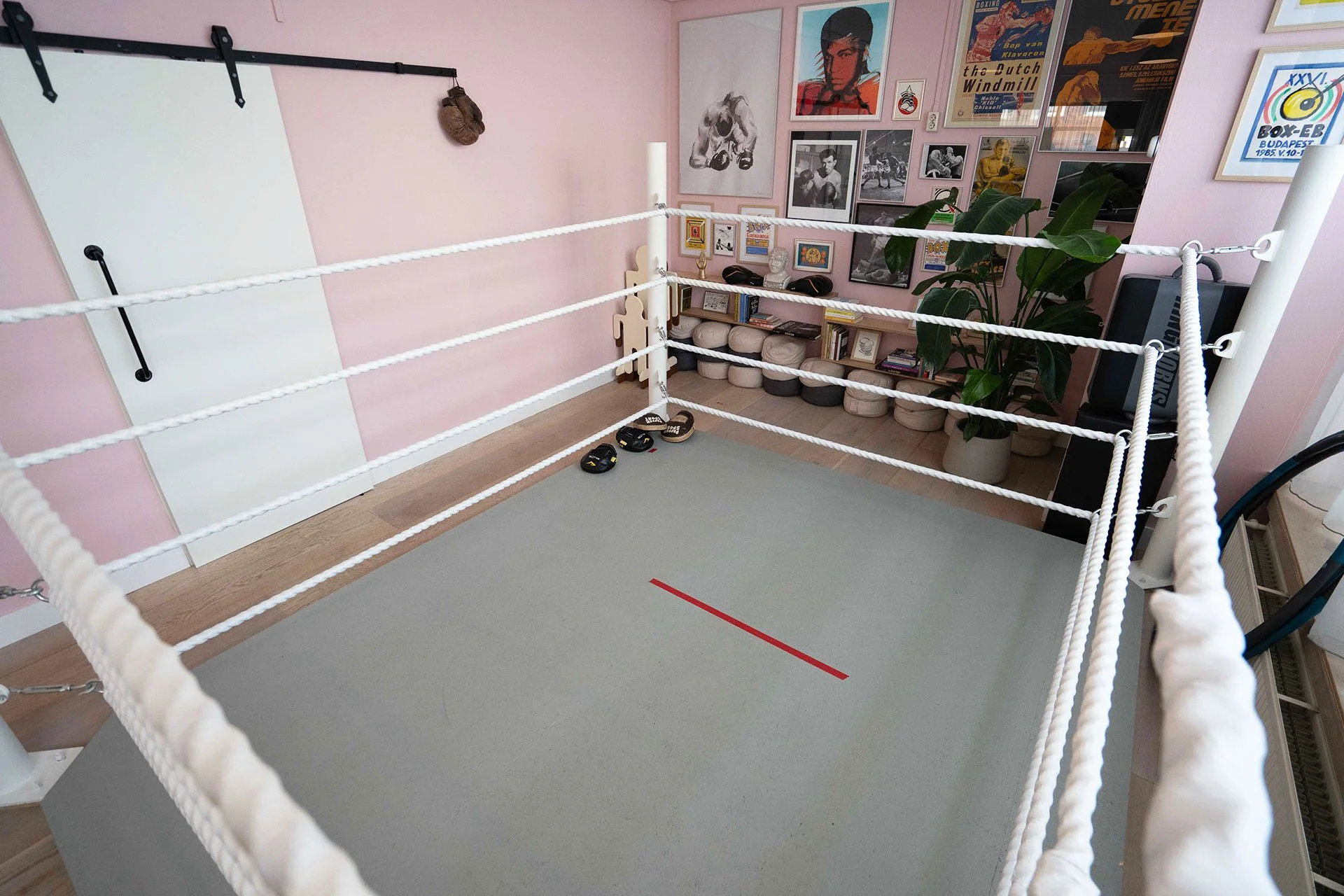 An empty indoor boxing ring with white ropes and a gray floor, situated in a room decorated with posters and artwork on pink walls.
