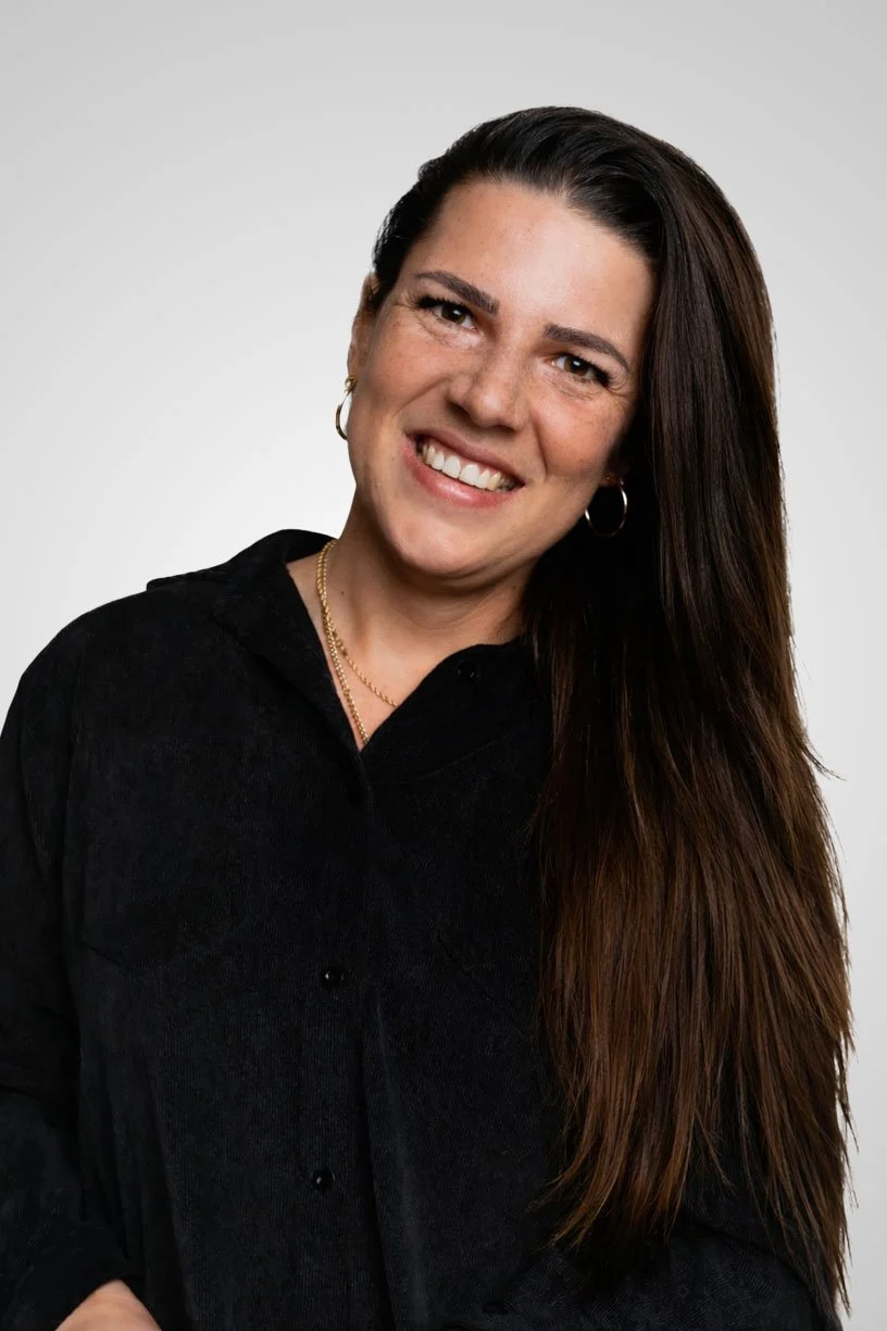 Portret van een vrouw met lange, donkere haar, lachend, gekleed in een zwarte blouse, met een witte achtergrond.