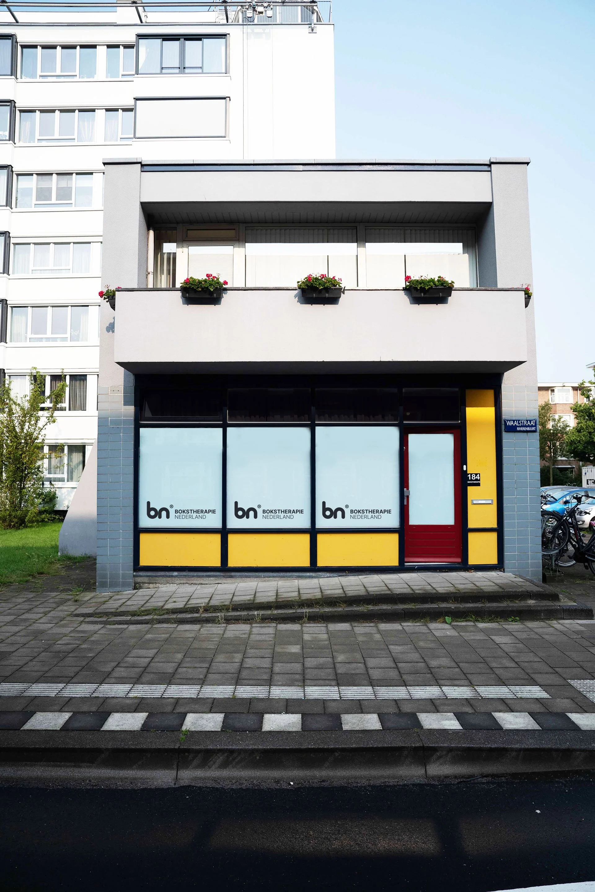 A two-story building with a ground-floor storefront featuring large glass windows and a red door. The windows display the logo and text 'bn BOKSTHERAPIE NEDERLAND.' The upper floor is a residential balcony with flower boxes. The building is located on a city street with a sidewalk and parked bicycles nearby.
