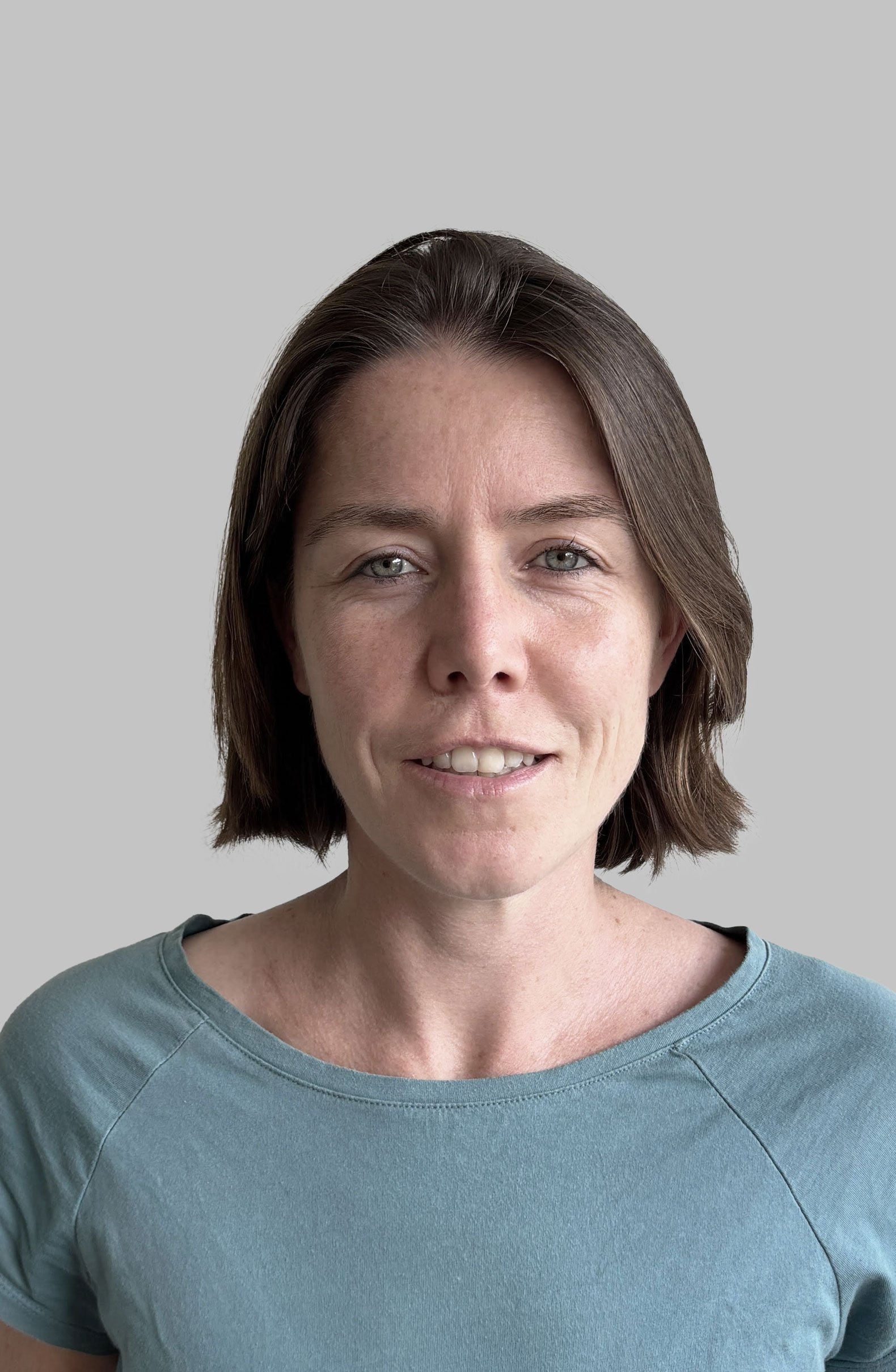 Close-up portrait of a woman with short brown hair, blue eyes, and fair skin wearing a light blue shirt, smiling slightly against a gray background.