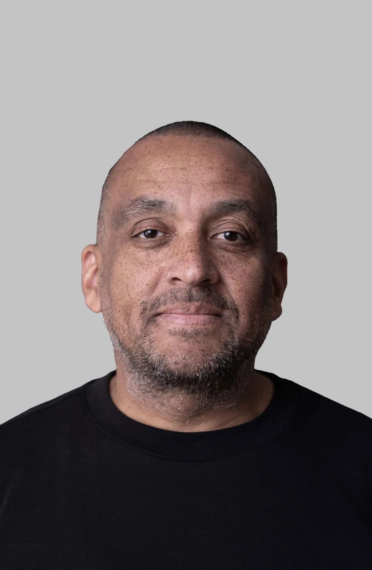 Close-up portrait of a middle-aged man with a beard and mustache, wearing a black shirt, against a neutral gray background.
