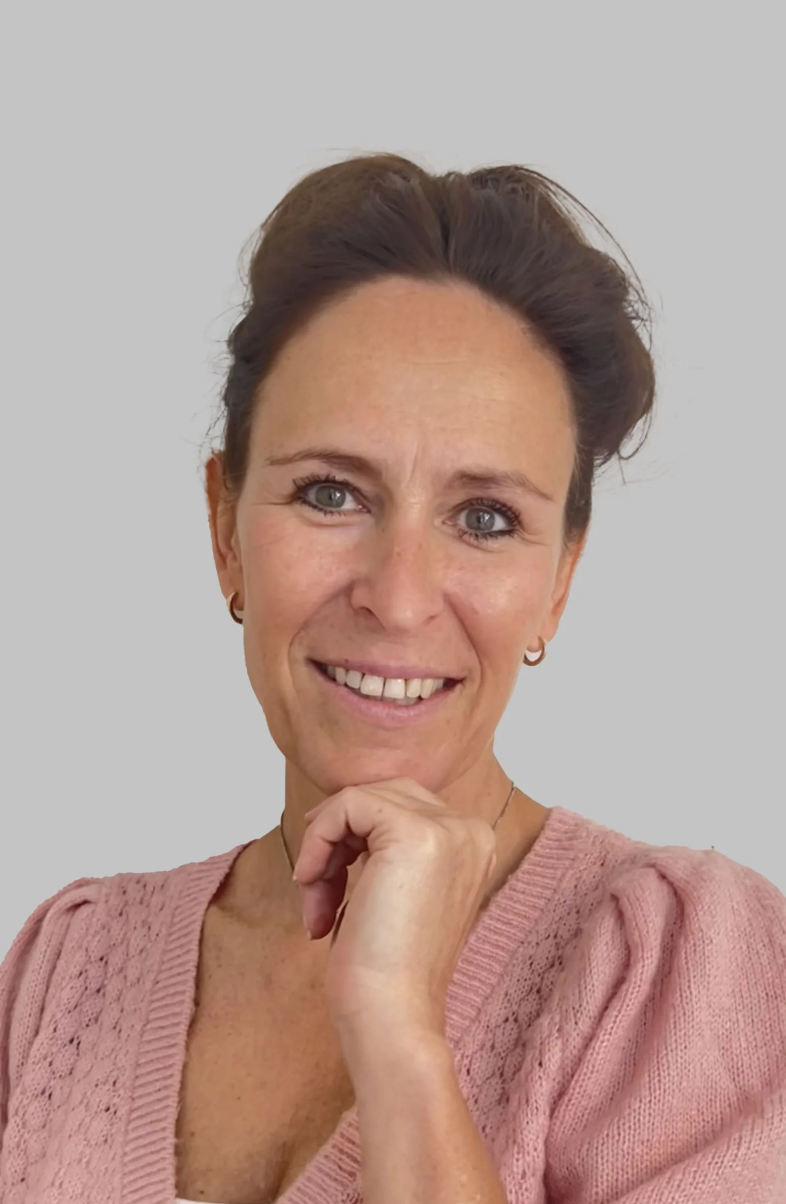 Close-up of a woman with short brown hair, blue eyes, and light skin, smiling and resting her chin on her hand, wearing a pink knitted top and small hoop earrings, against a plain light gray background.