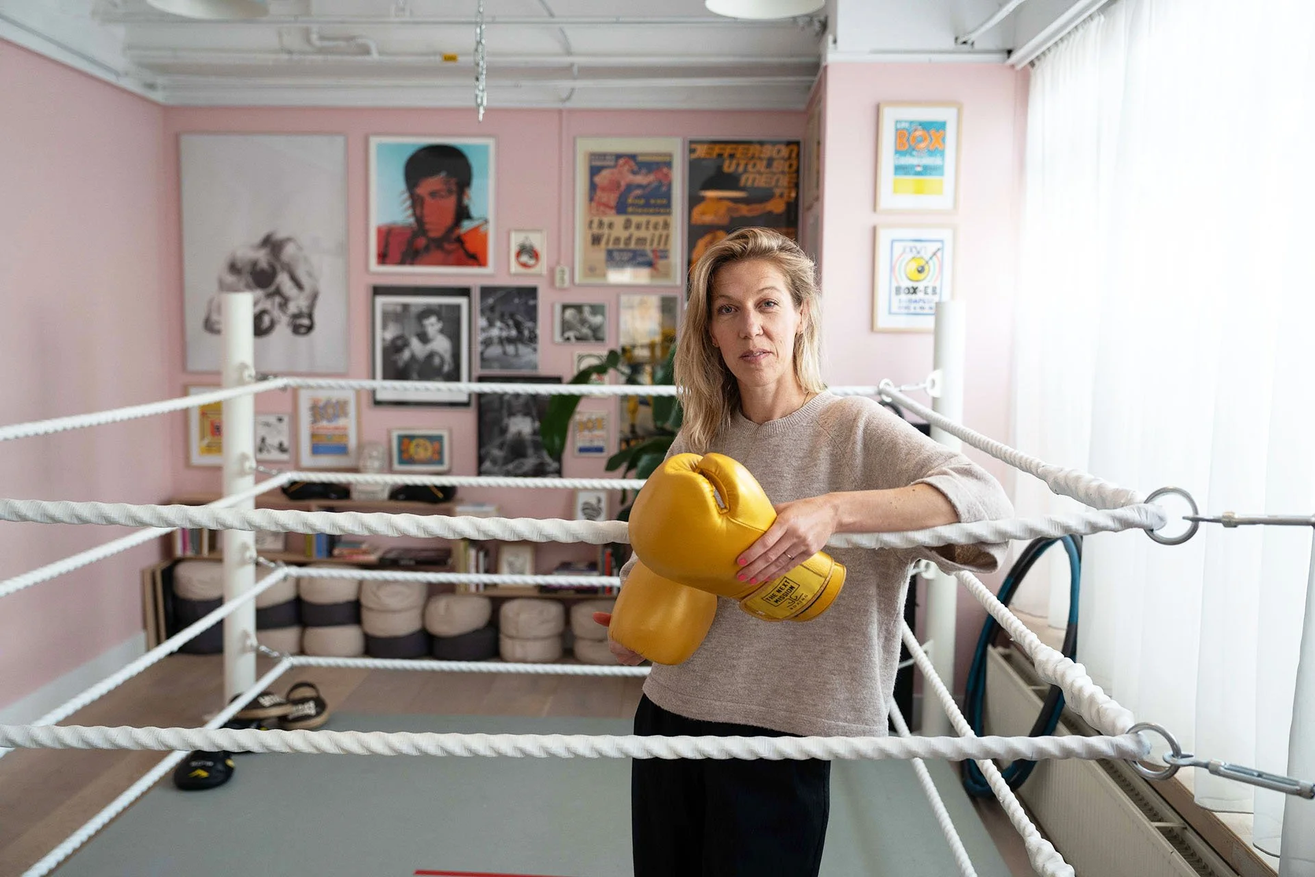 A woman in a boxing ring wearing yellow boxing gloves, standing inside the ropes with her hands resting on them, in a room with pink walls and boxing posters.