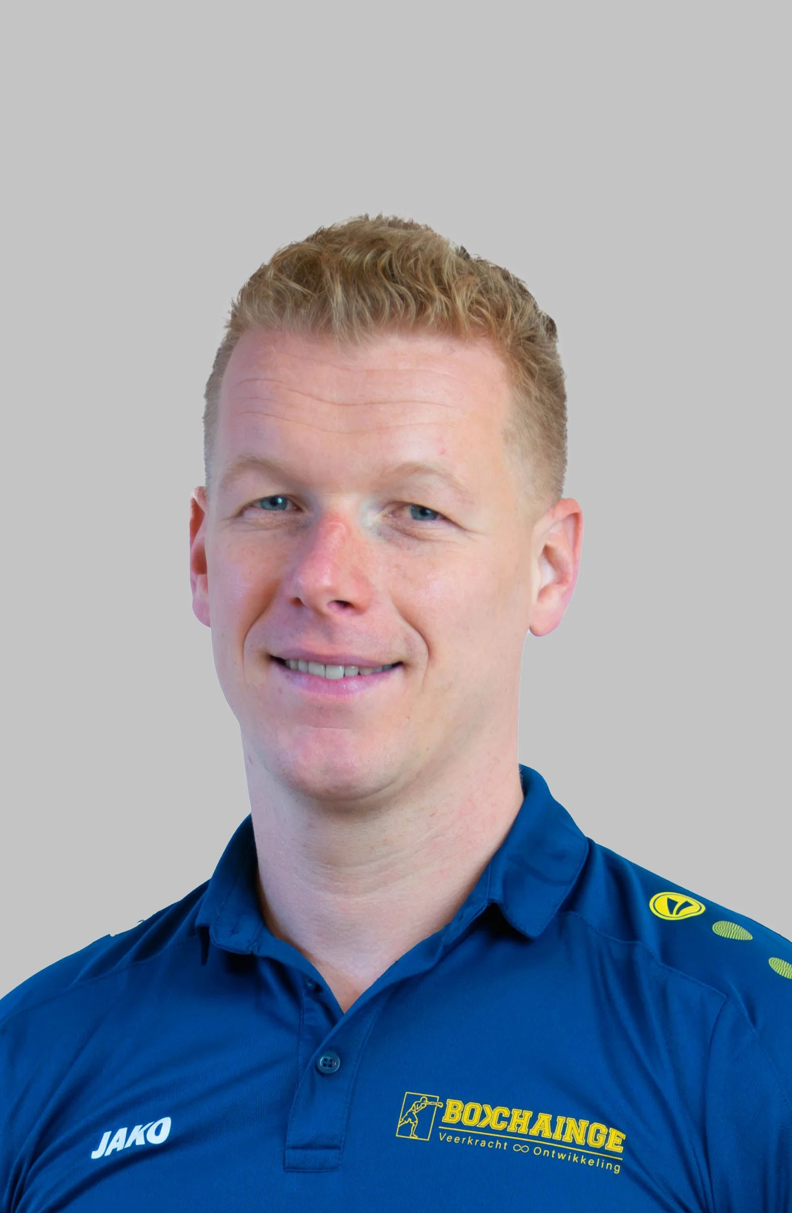 Portrait of a man with short, curly red hair, wearing a blue sports shirt with a logo on the chest, smiling at the camera.