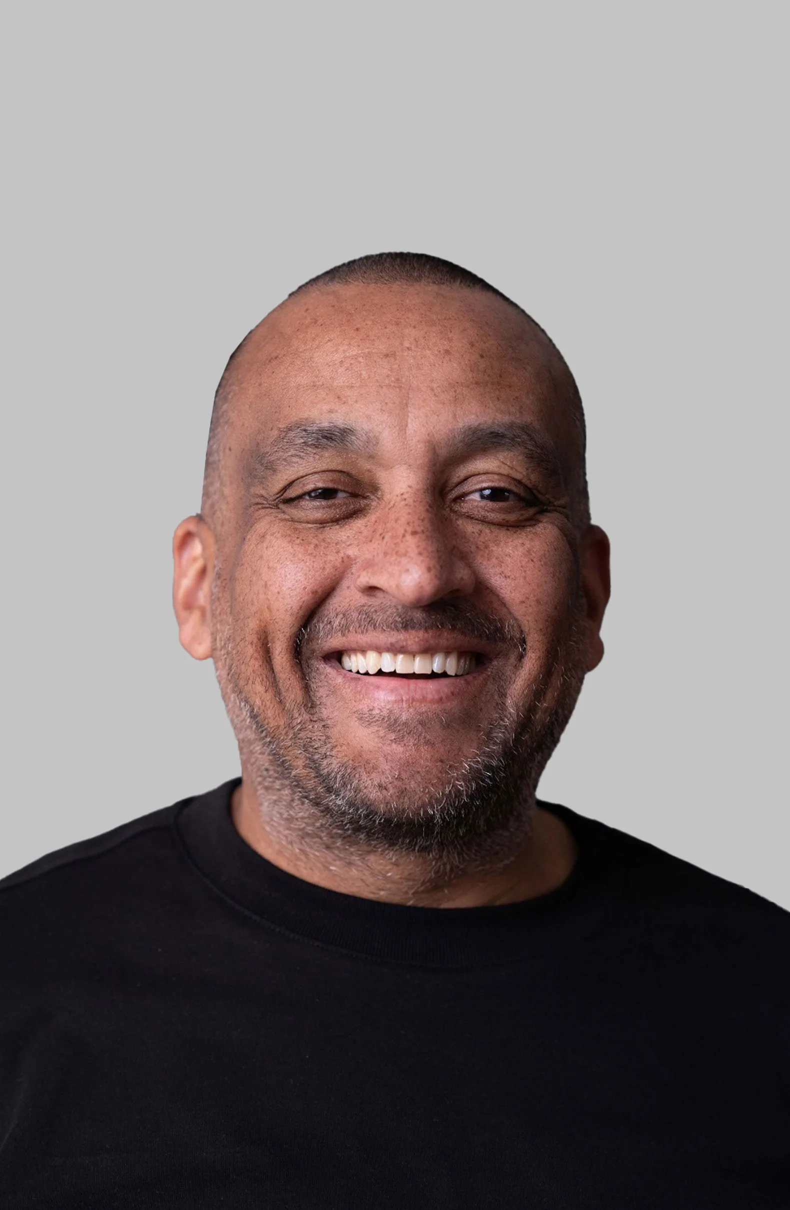 Close-up of a smiling man with a bald head, facial hair, and freckles, wearing a black shirt against a plain gray background.