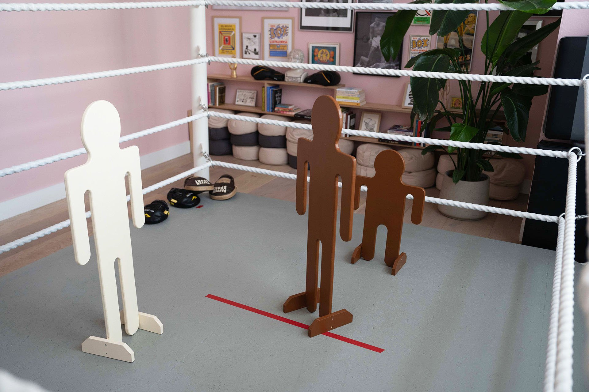 A boxing ring with wooden cutouts of two fighters inside, lined with white ropes, and a red line on the gray floor indicating the center.
