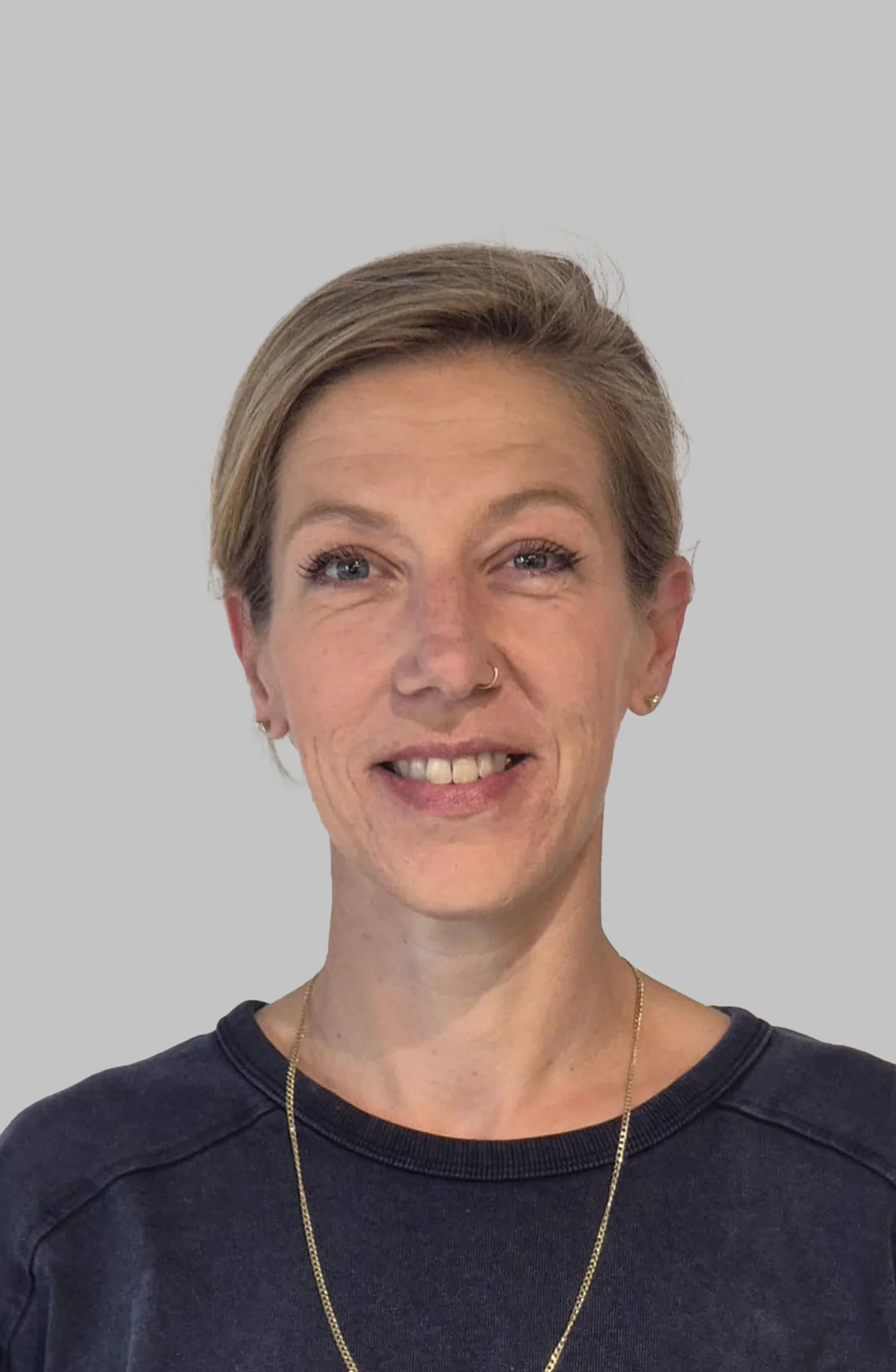 A portrait of a smiling woman with short light brown hair, blue eyes, a nose ring, and earrings, wearing a black shirt and a gold necklace, against a plain light gray background.
