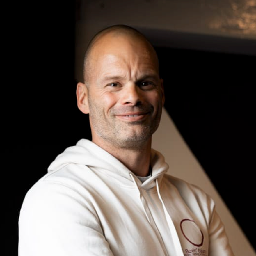 Man met korte baard en korte haar, glimlachend, draagt een witte hoodie, binnenruimte met een donkere achtergrond.