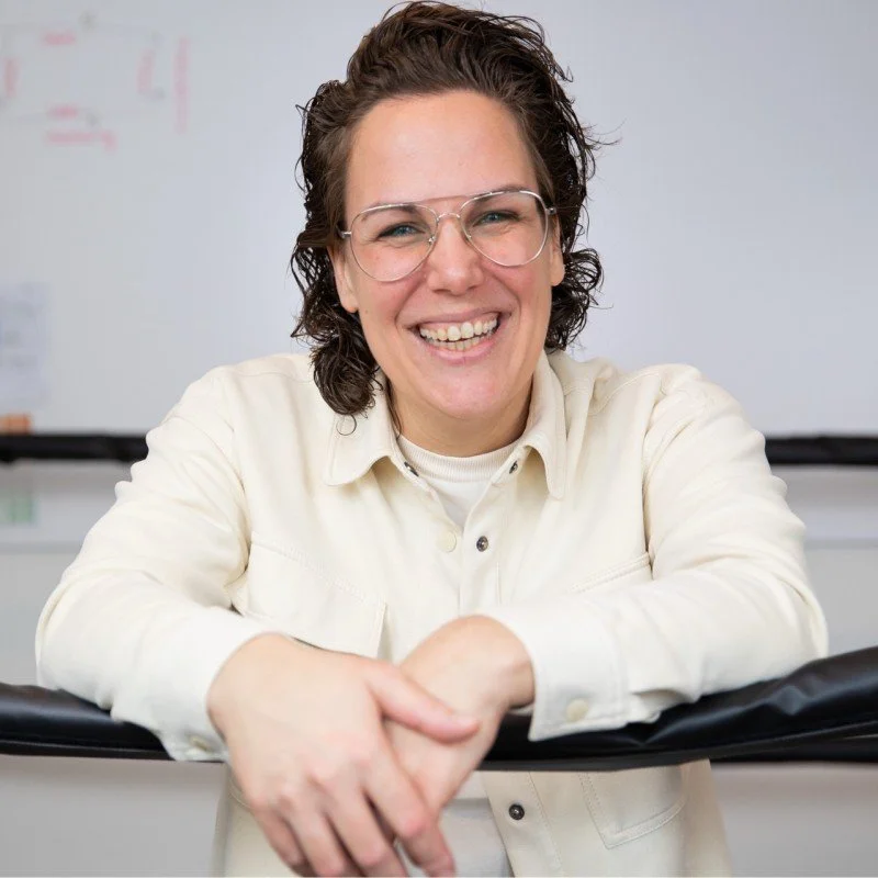 Een lachende jonge vrouw met brillen, gezeten in een klaslokaal, die naar de camera kijkt.