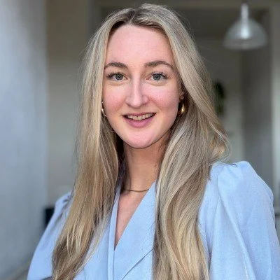 Een vrouw met lang blond haar, glimlachend en gekleed in een lichtblauwe blouse, voor een lichte achtergrond.