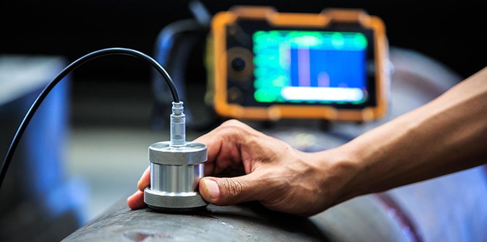 A person's hand holding a metallic sensor connected by a wire to an electronic testing device, with a digital screen in the background displaying waveforms.