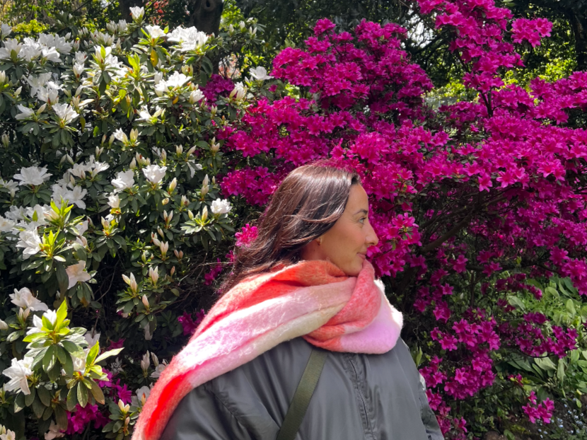 Mikkenzie with long dark hair standing in front of blooming pink and white flowers with green foliage, wearing a vibrant pink, coral and white skarf and khaki coloured jacket.