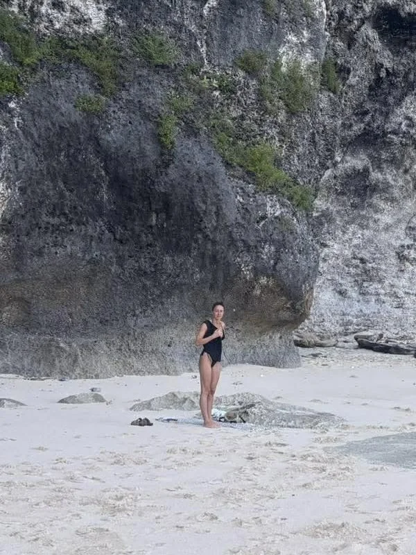 Mikkenzie in a black swimsuit standing far in the distance on a beach with a sandy shore, rocks, and a large rocky cliff in the background.