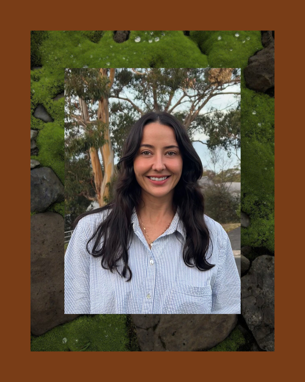 Mikkenzie smiling with long dark hair, wearing a blue and white striped shirt, standing outdoors with trees in the background.