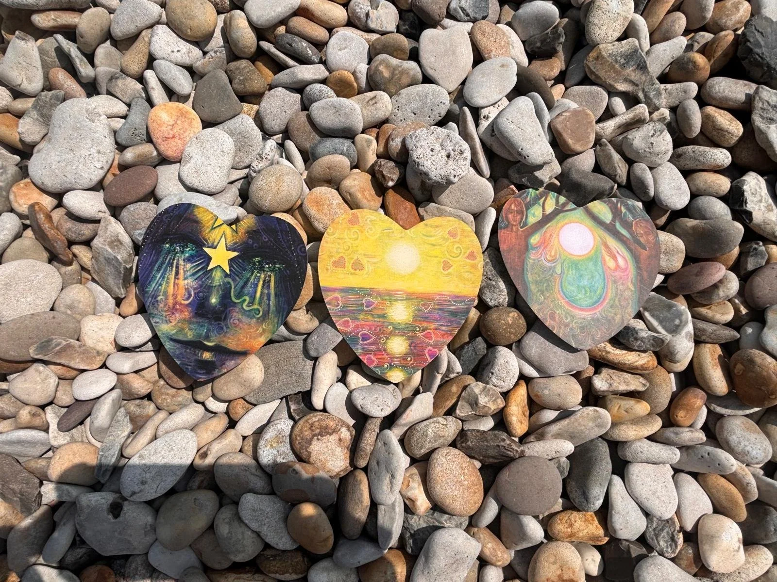 Three colorful, decorated heart-shaped paper cutouts are lying on a bed of small, multicolored pebbles.
