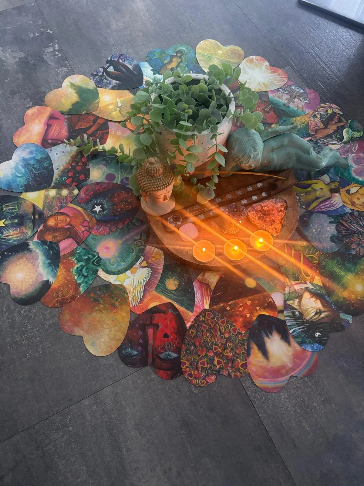 A collection of colorful paper hearts featuring various artistic designs and images, arranged in a circle around a pot of green succulent plants. On a wooden surface in the center, there are small decorative sculptures, a glow of three lit candles, and a couple of small rocks.