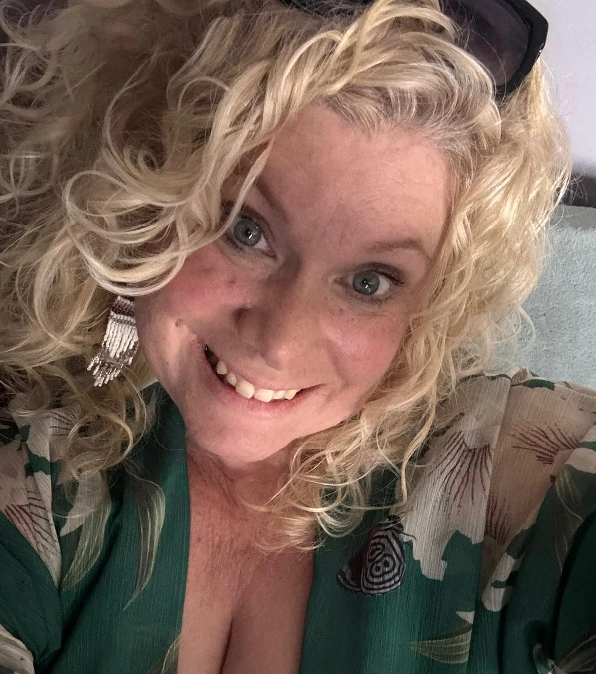 A woman with curly blonde hair smiling at the camera, wearing a patterned blouse and large earrings.