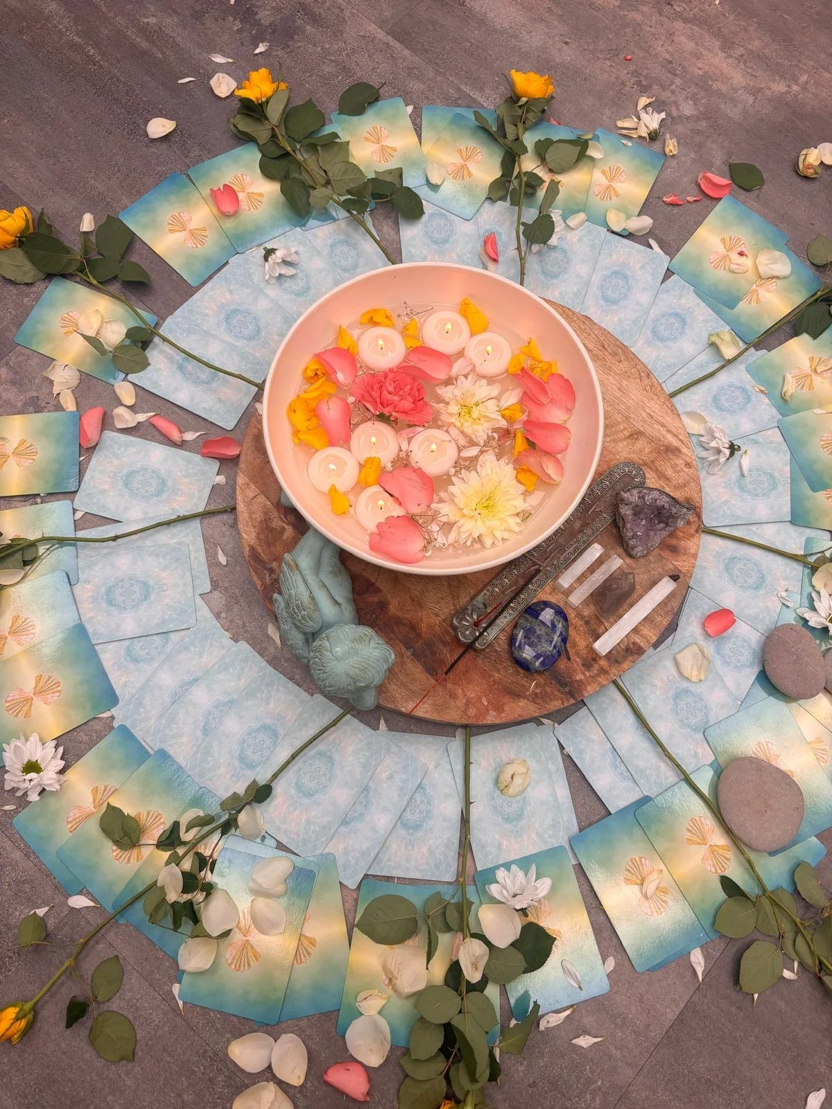 A spiritual or ceremonial altar arranged with candles, flowers, stones, and tarot or oracle cards, featuring a central bowl with floating candles and flower petals, surrounded by white daisies, rose petals, and eucalyptus leaves on a circular wooden surface.