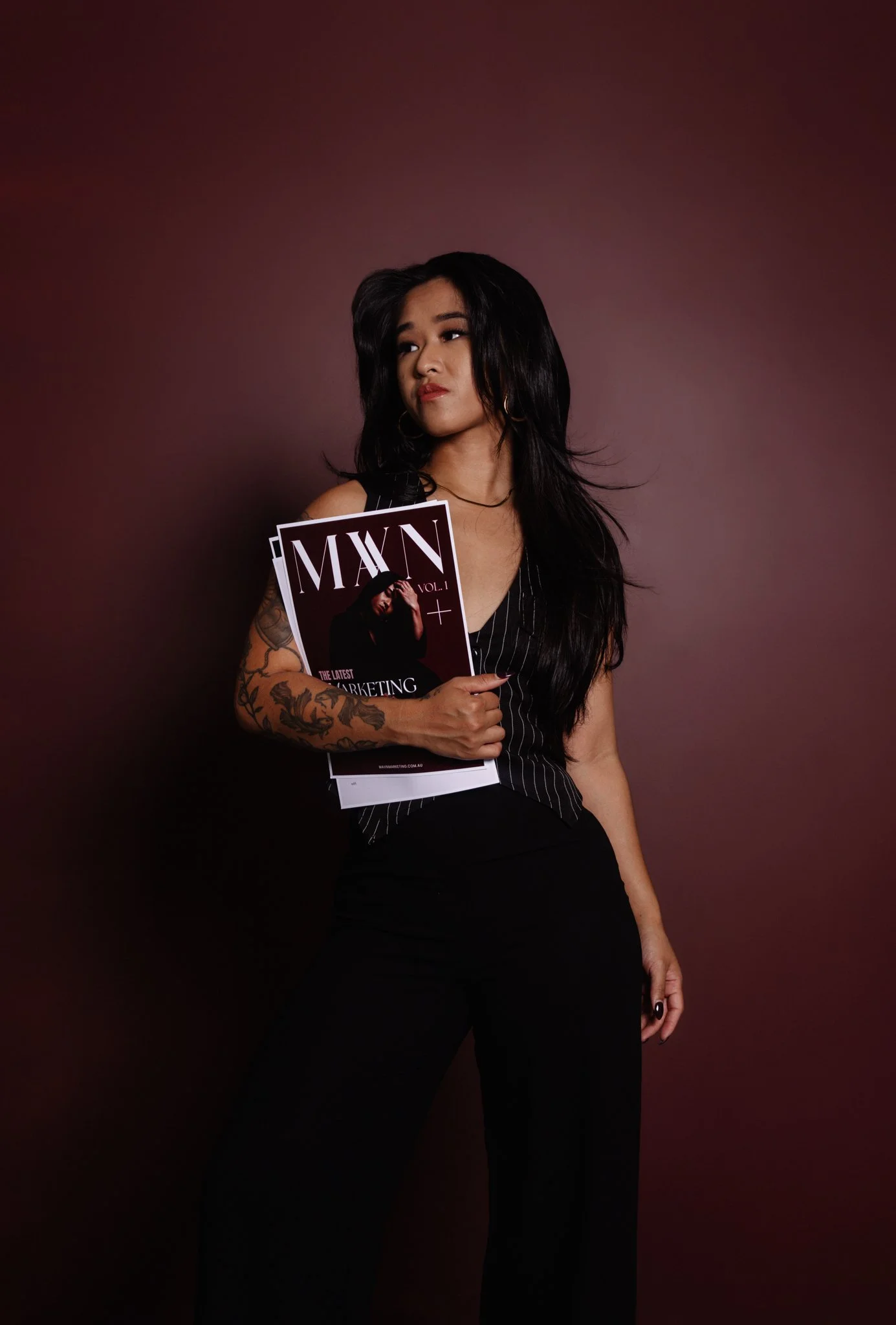 A woman with long dark hair and tattoos on her left arm is holding a magazine titled "MAXN" with a picture of a woman on the cover. She is wearing a black pinstripe top and black pants, standing against a dark maroon background.