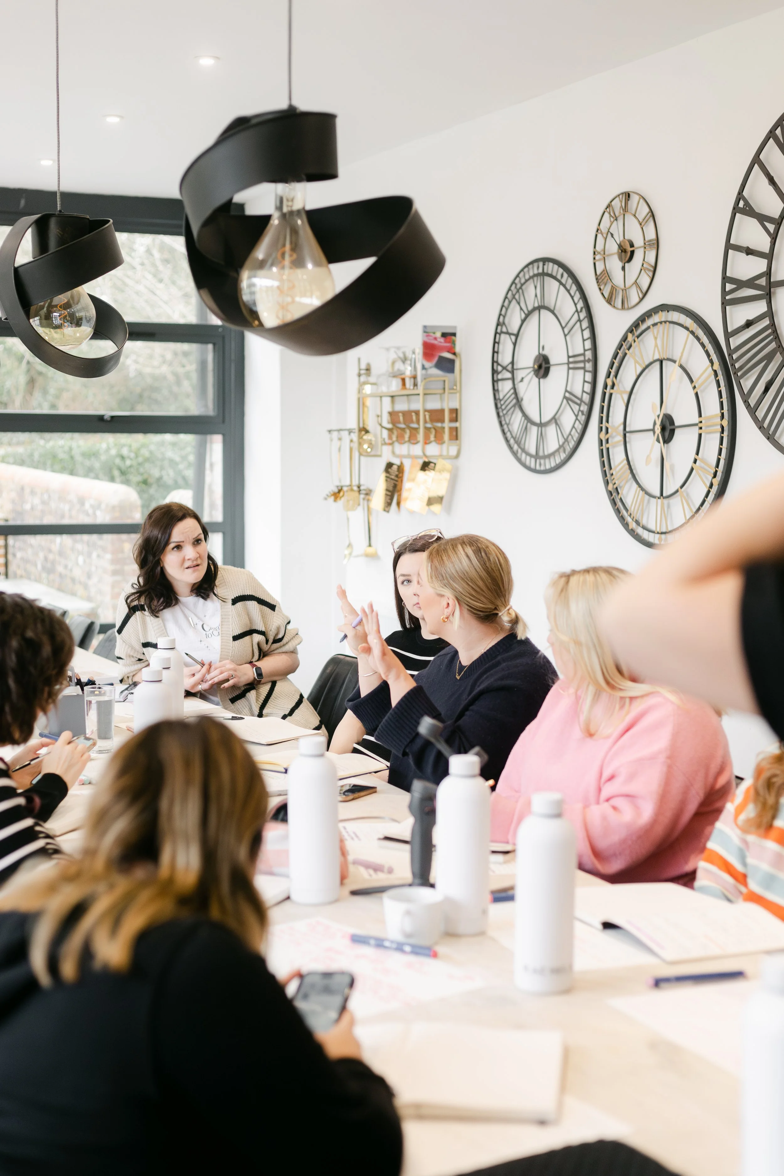 A group of women participating in a meeting or discussion around a conference table in a modern room with large wall clocks and decorative lighting.